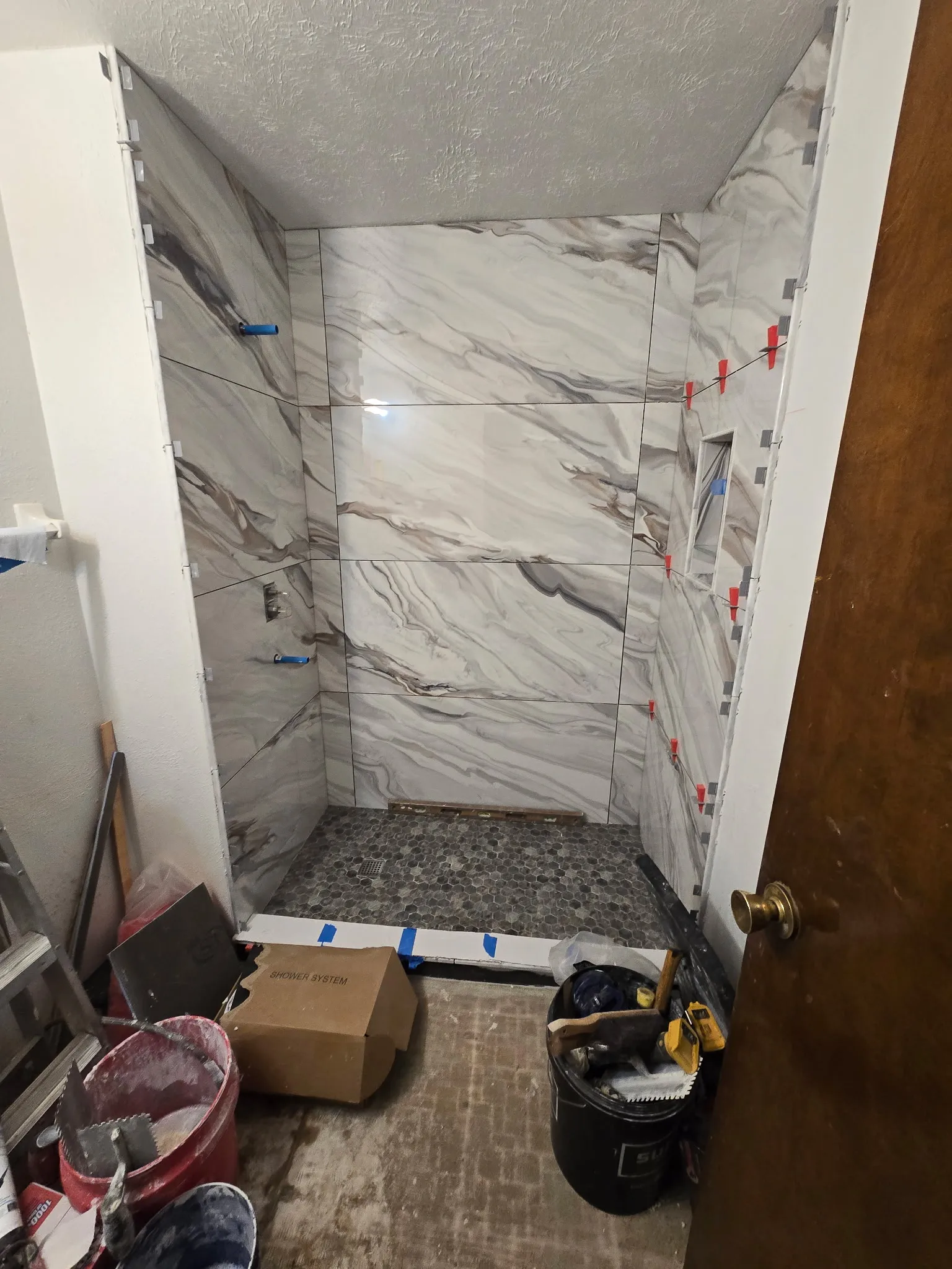 Marble-look porcelain shower walls part-tiled with levelling clips still in place, over a pebble mosaic pan